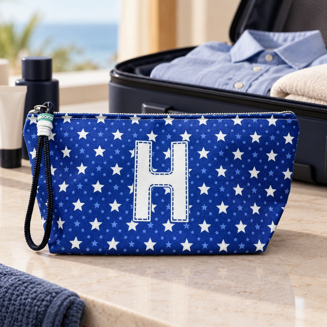 Bolsa Alex azul com letra H fechada num cenário de viagem com mala, roupa e artigos de higiene masculina.