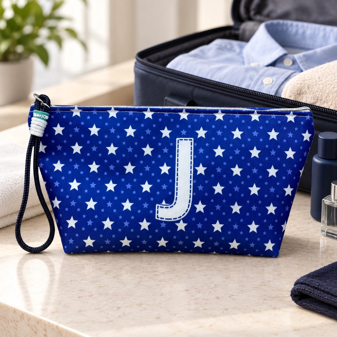 Bolsa Alex azul com letra J fechada num cenário de viagem com mala, roupa e artigos de higiene masculina.