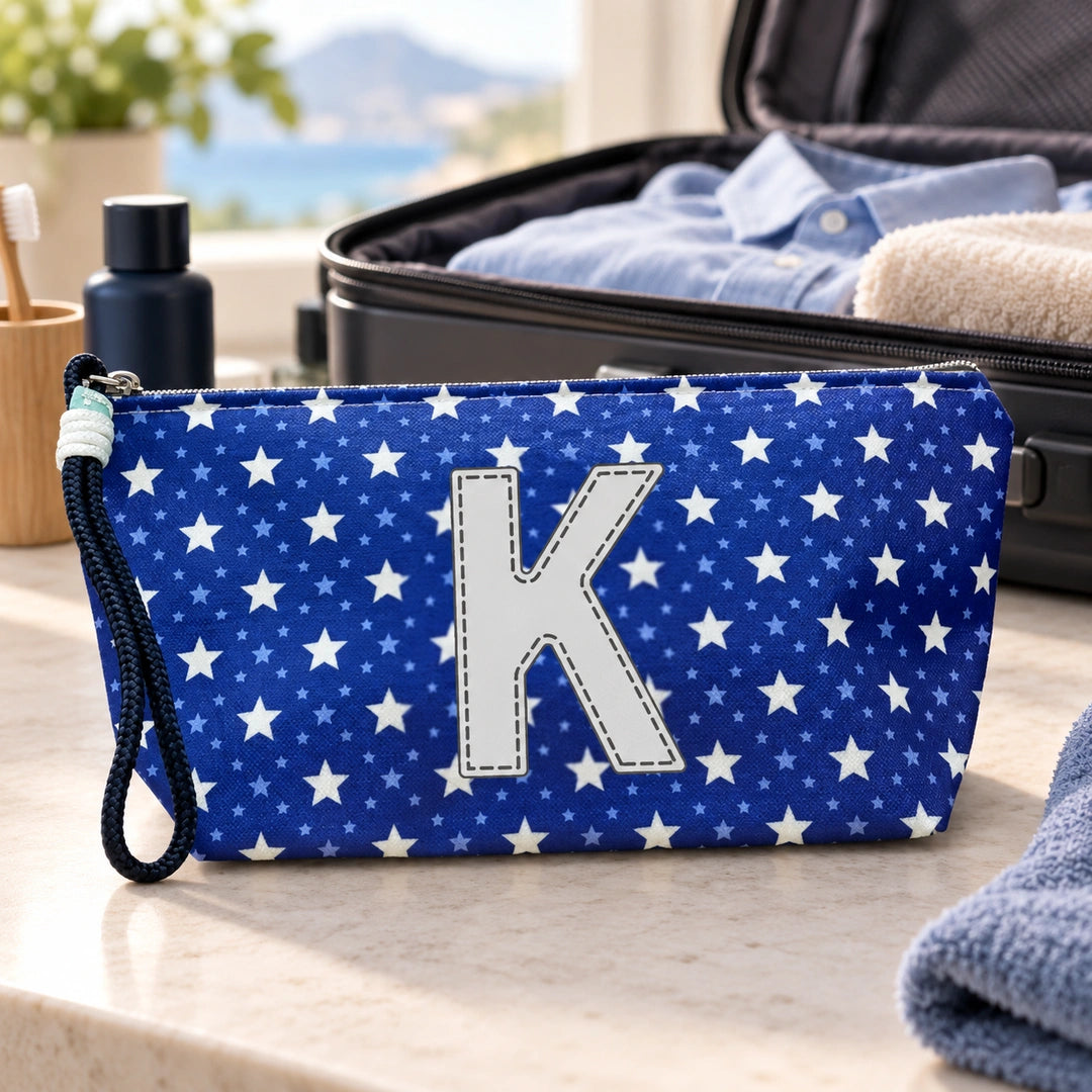 Bolsa Alex azul com letra K fechada num cenário de viagem com mala, roupa e artigos de higiene masculina.
