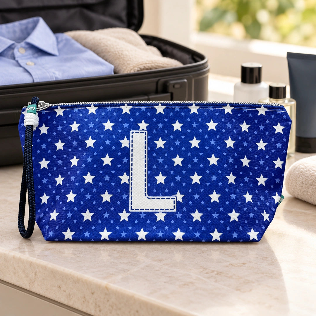 Bolsa Alex azul com letra L fechada num cenário de viagem com mala, roupa e artigos de higiene masculina.
