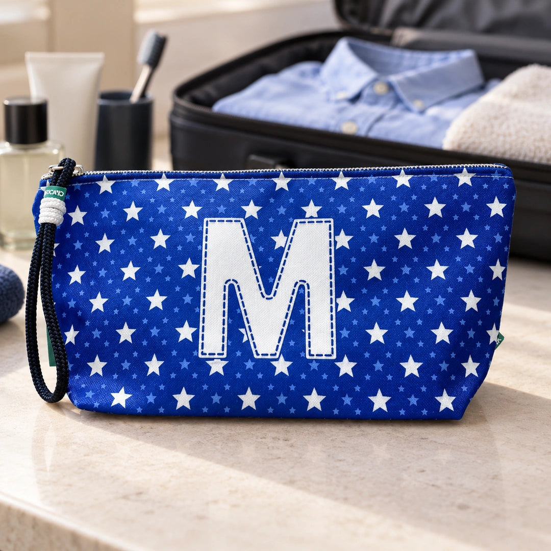 Bolsa Alex azul com letra M fechada num cenário de viagem com mala, roupa e artigos de higiene masculina.