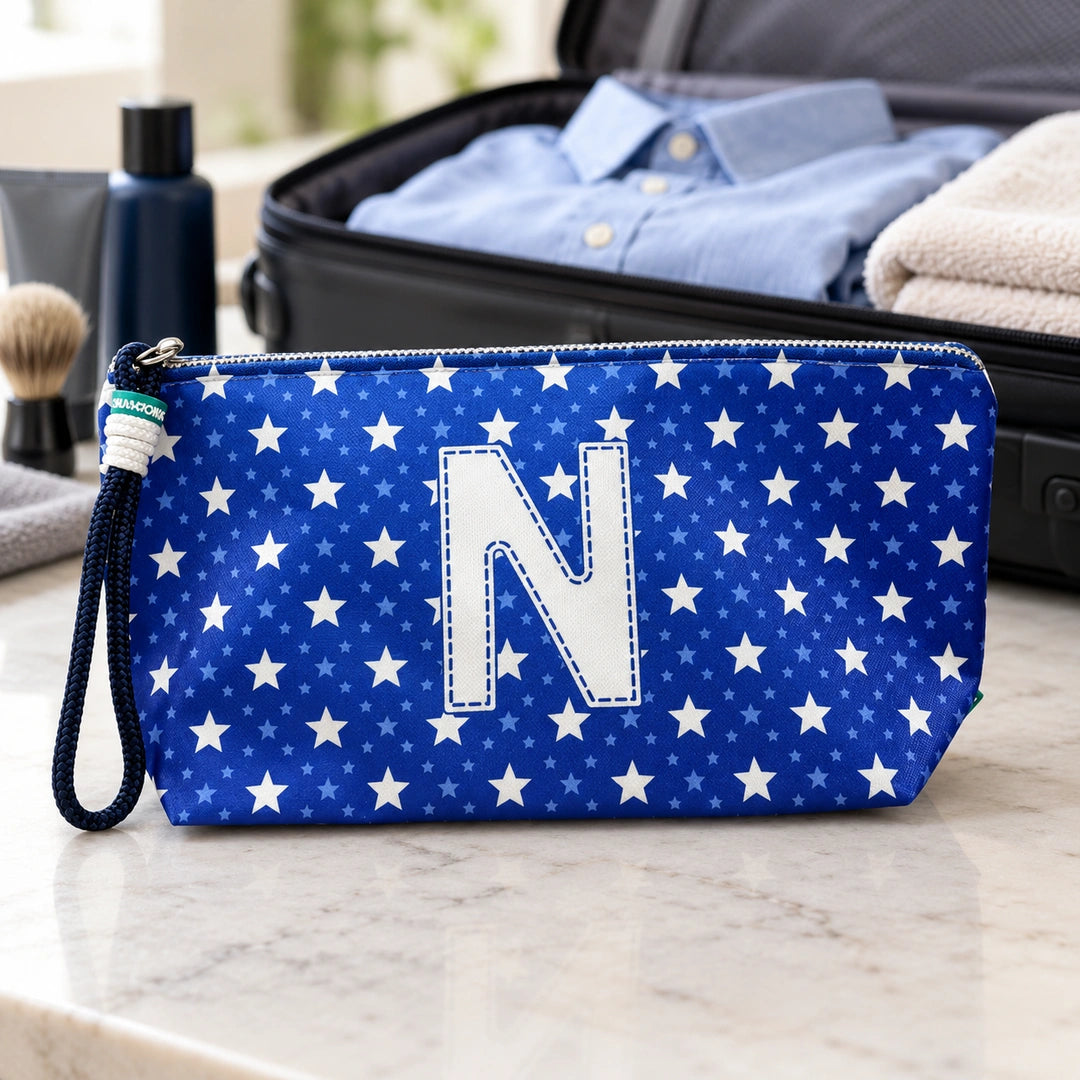 Bolsa Alex azul com letra N fechada num cenário de viagem com mala, roupa e artigos de higiene masculina.
